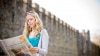 A young woman with blonde hair reads a newspaper in front of a large building