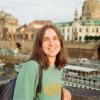 A young woman smiling while standing on a bridge with historic buildings and a river in the background on a sunny day in Germany.